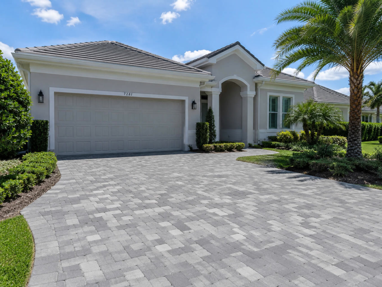 Gray tumbled paver driveway curving up to the two car garage for a Jupiter homeowner, framed by clipped hedges and palms