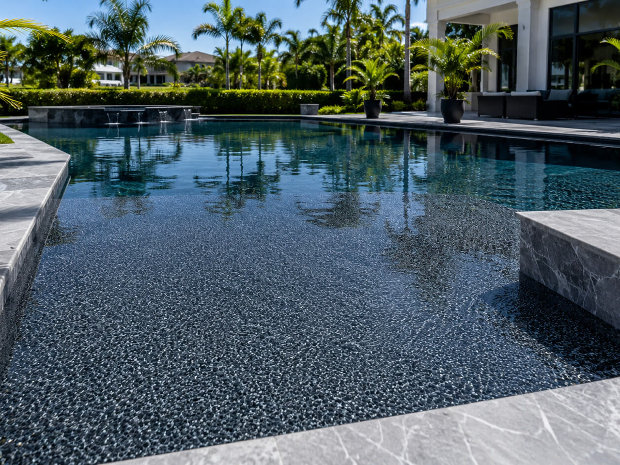 Dark charcoal pebble pool finish under still water, framed by gray marble coping and spillway at a Boynton Beach pool