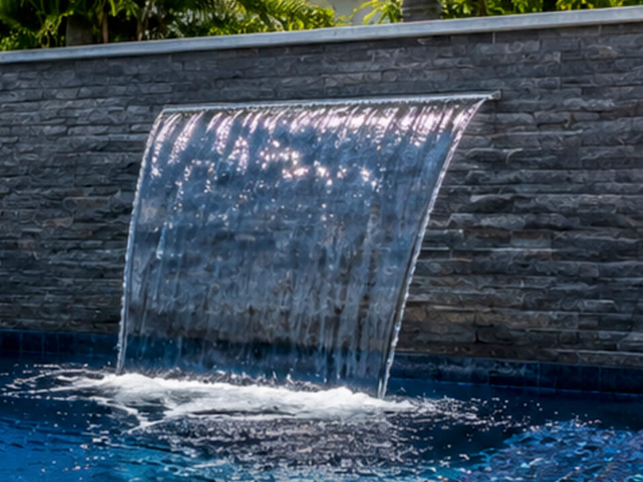 Sheer descent waterfall blade falling from a gray stacked stone wall into a deep blue Delray Beach pool
