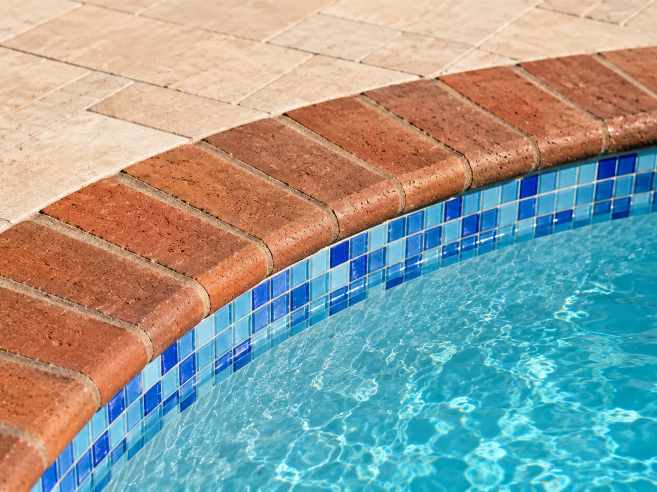 Red brick coping following the curve of a pool over a blue mosaic waterline tile, beside a travertine deck at a Jupiter residence